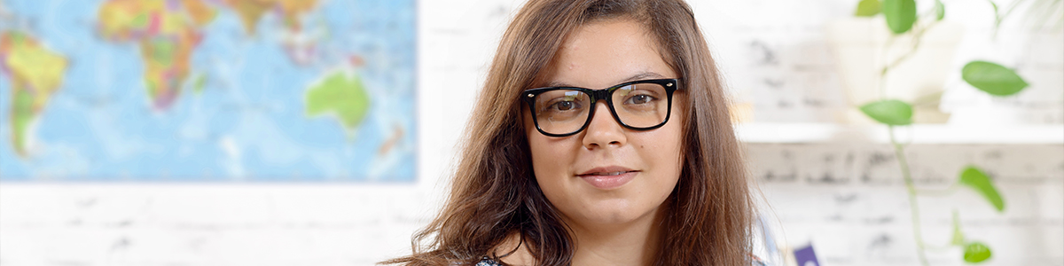 A woman with long brown hair and black glasses is shown in front of a world map and a white wall with green plant leaves.