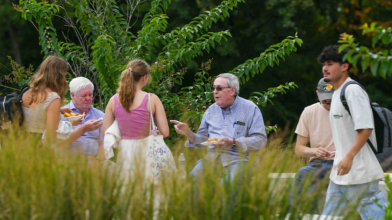 A group of people gathered outdoors, enjoying food and conversation. Two older men are seated, while three younger individuals stand nearby.