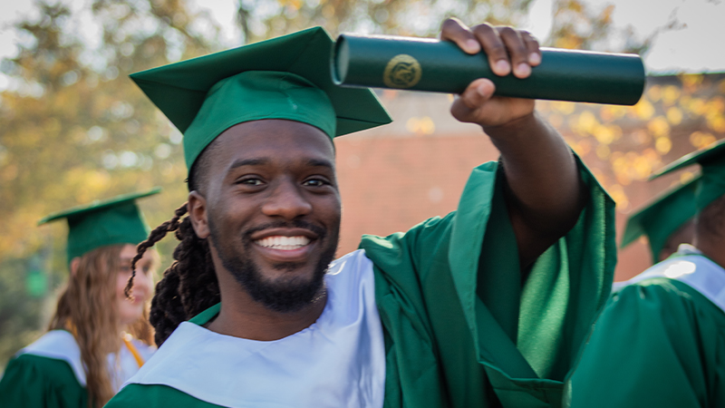 A smiling graduate wearing a green cap and gown holds up a diploma in one hand, surrounded by fellow graduates in the background.
