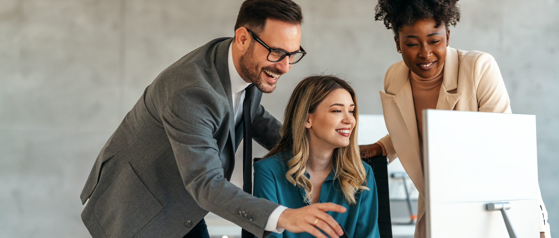 Three colleagues gather around a computer in a modern office. A man in a suit points at the screen while two women watch, one smiling.