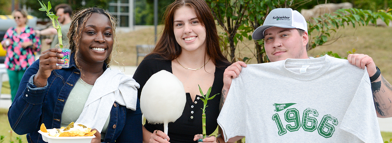 Three young adults pose outdoors at a fair, holding a cotton candy, a snack tray, and a white T-shirt printed with 1966.