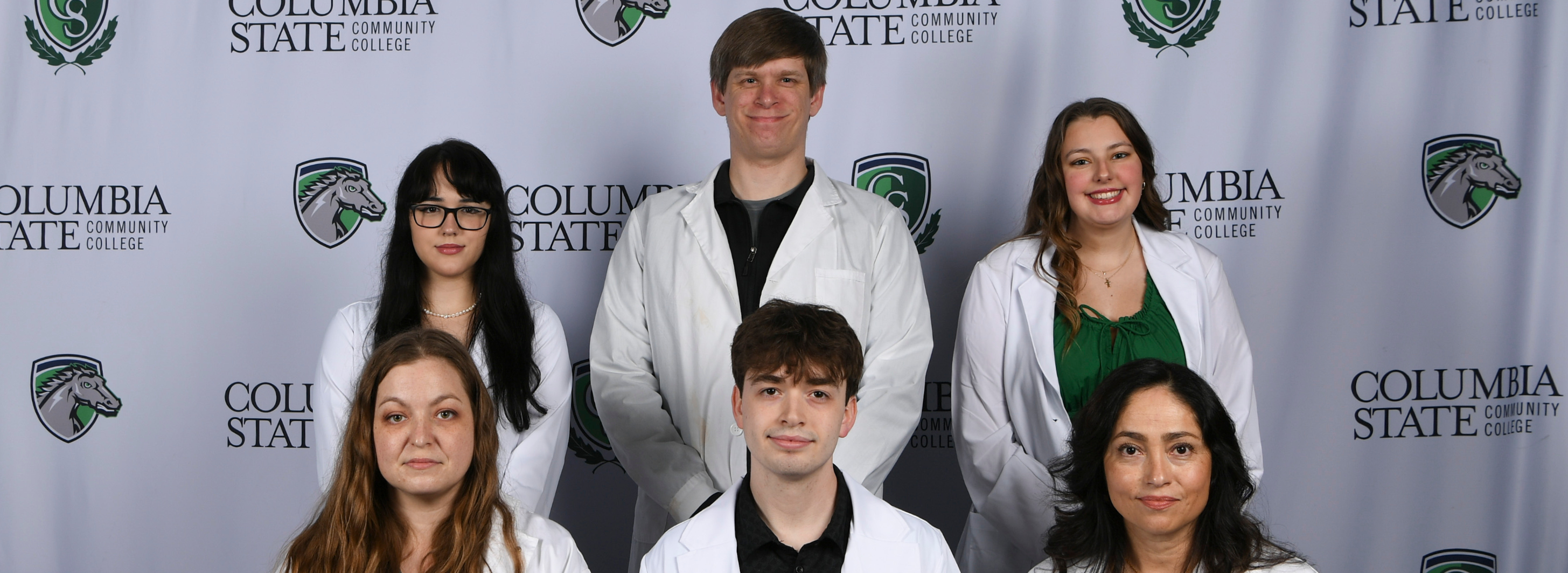 A group of six people sit and stand for a photo in front of a white background.