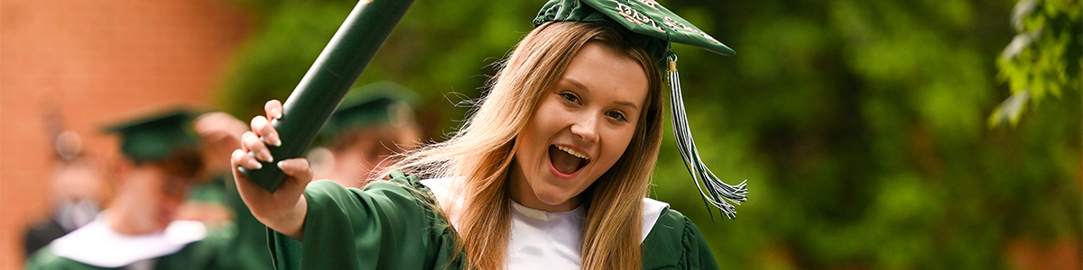 A smiling graduate in a green cap and gown holds up a diploma tube outdoors, with other graduates blurred in the background.