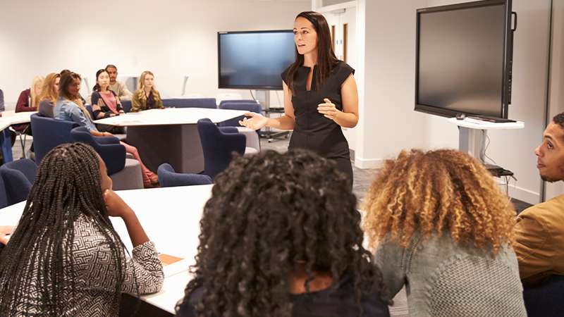 A woman in a black dress speaks to a group seated around tables in a modern conference room with wall-mounted screens.