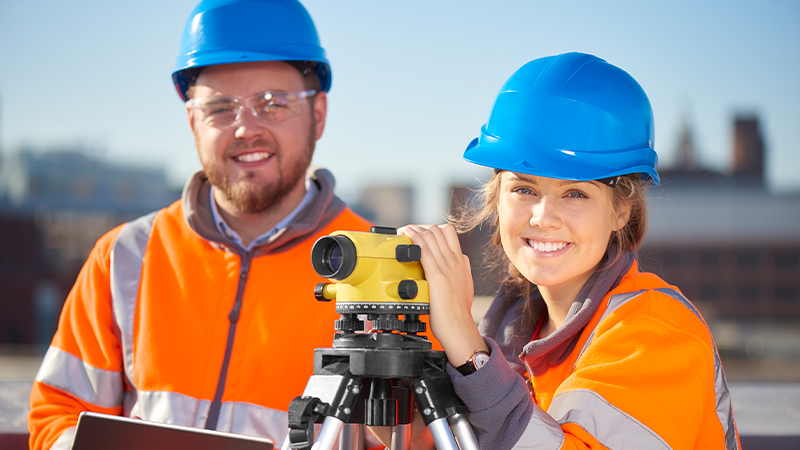 A woman and a man wearing blue hard hats and orange safety vests stand beside a yellow surveying instrument on a tripod, with buildings blurred in the background.
