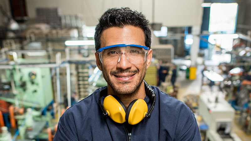 A man wearing blue safety glasses and yellow earmuffs stands in a factory, with industrial machinery and workstations blurred in the background.
