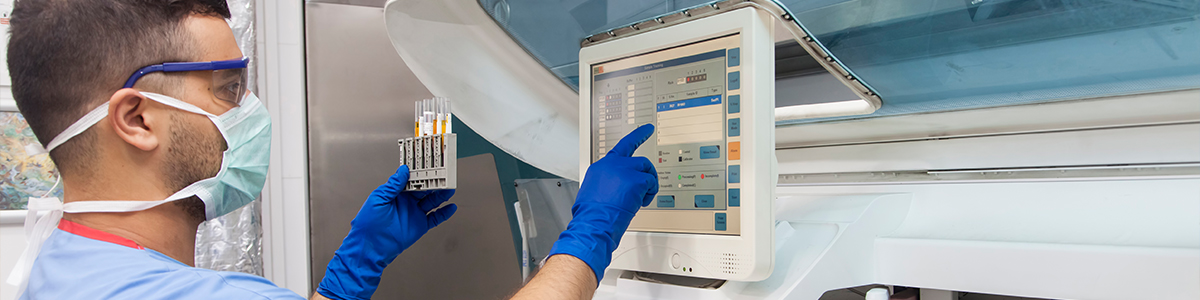 A masked technician in blue gloves points to a touchscreen control panel beside laboratory equipment and holding test tubes.